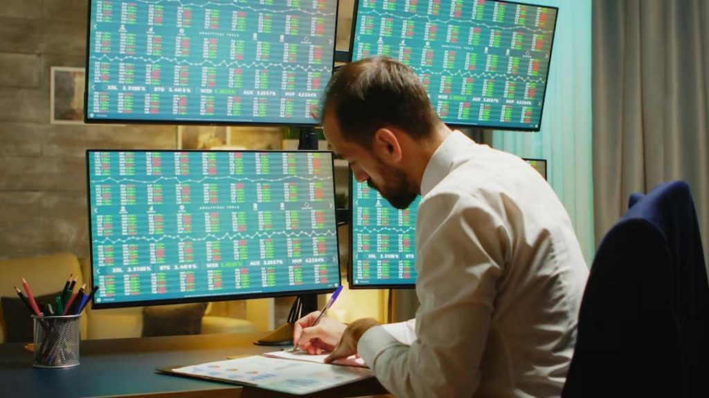A man sitting in front of four monitors with stock charts, writing trading signals on a sheet of paper.