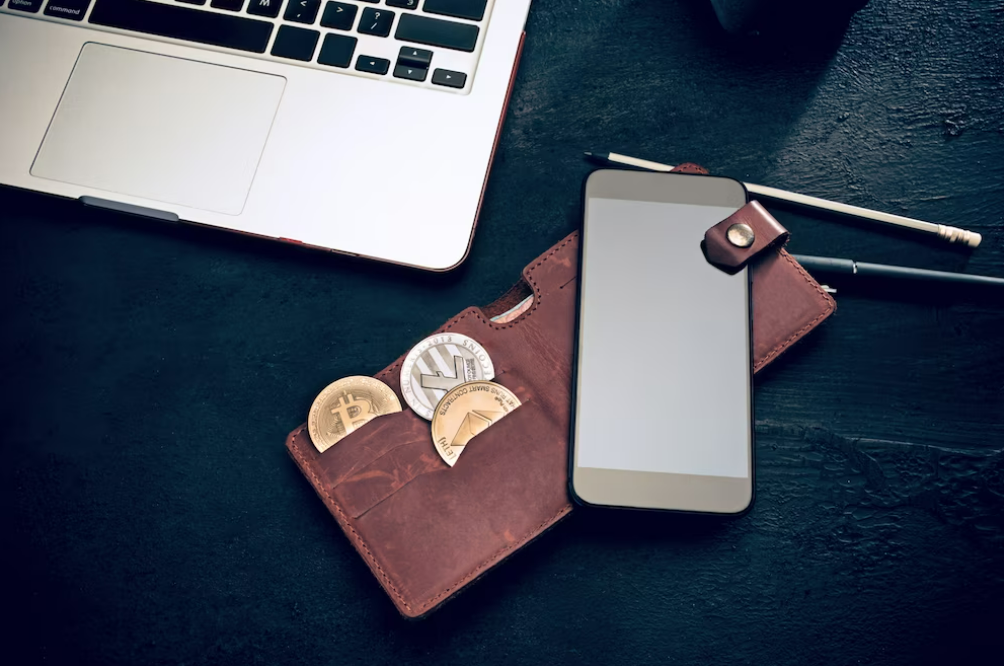 Teléfono móvil y cartera con representaciones de criptomonedas, sobre fondo oscuro y vista parcial de computadora portátil.