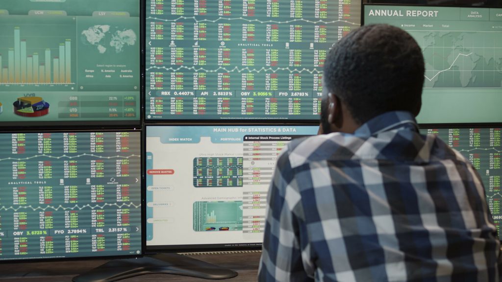Un hombre de espaldas mirando seis monitores con gráficos de acciones.
