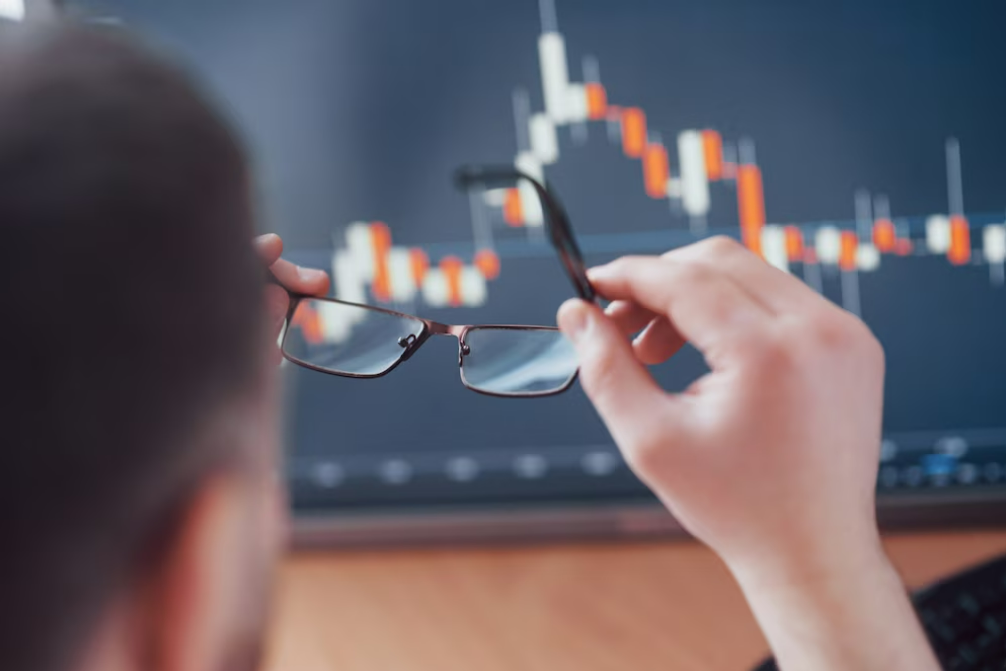 Hombre de espaldas sosteniendo sus gafas mientras mira una pantalla con gráficos de velas japonesas